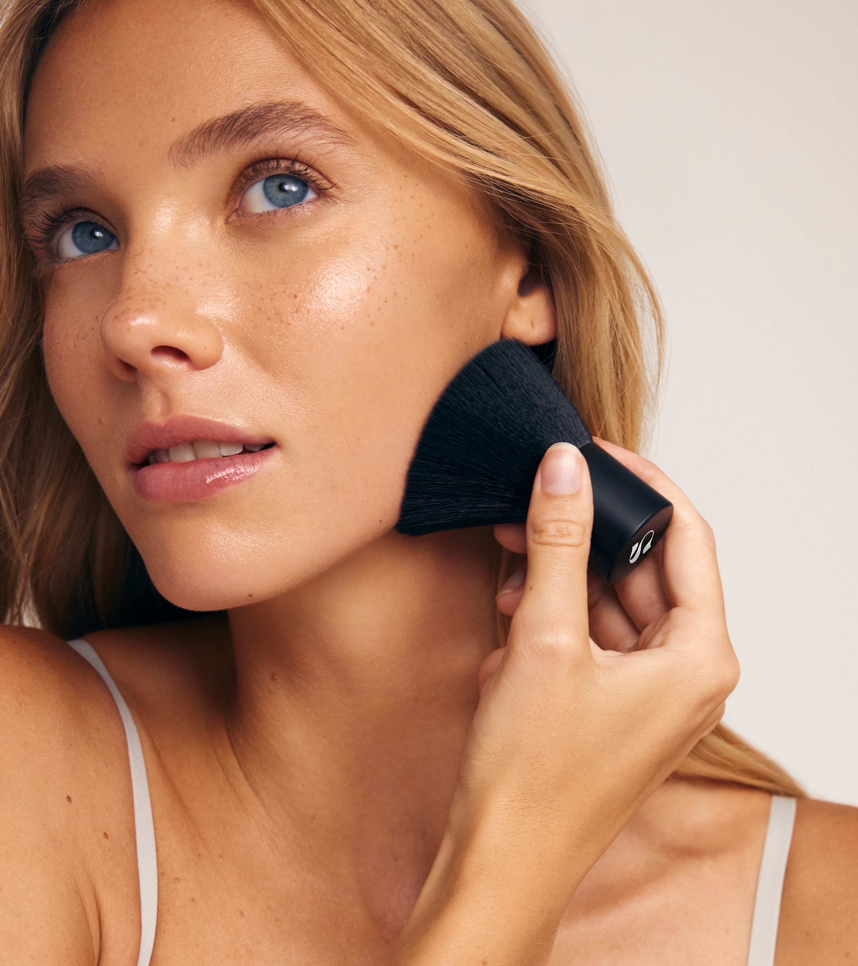 A woman with light skin, blue eyes, and long blonde hair uses The Face Set’s large face brush on her cheek, perfecting a flawless face tan while gazing upward in a light gray strap top against a neutral beige background.