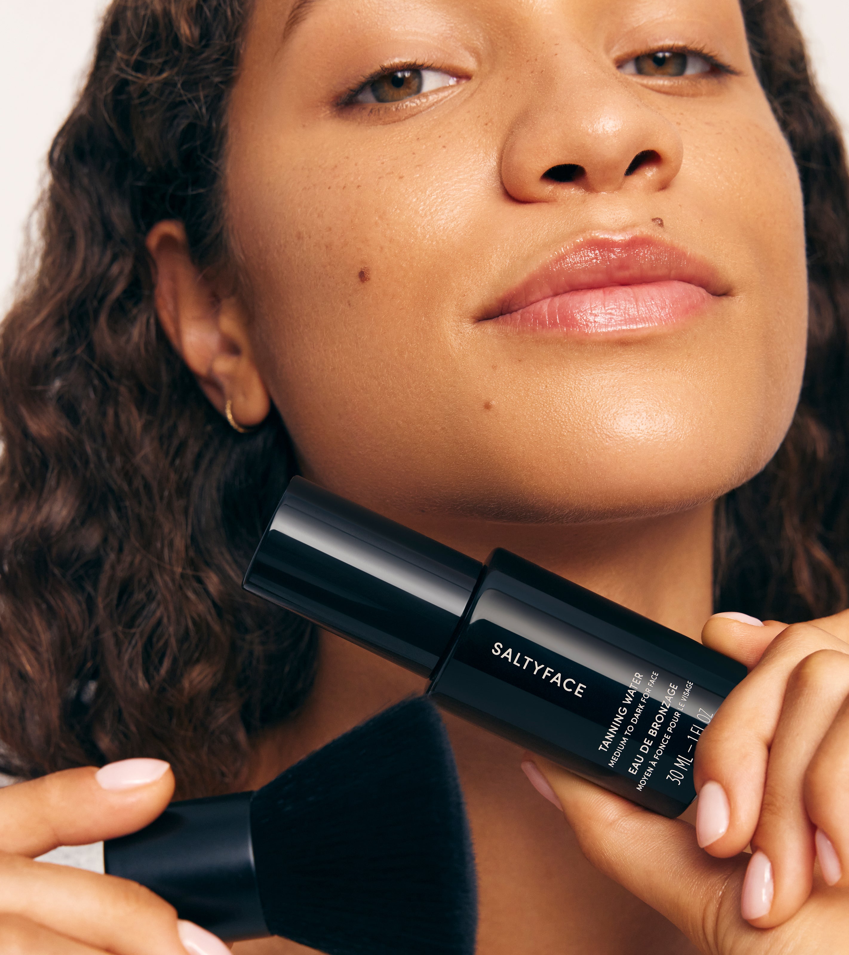A woman with curly hair holds The Face Set bottle and a makeup brush near her face, showcasing smooth, glowing skin and a confident expression.
