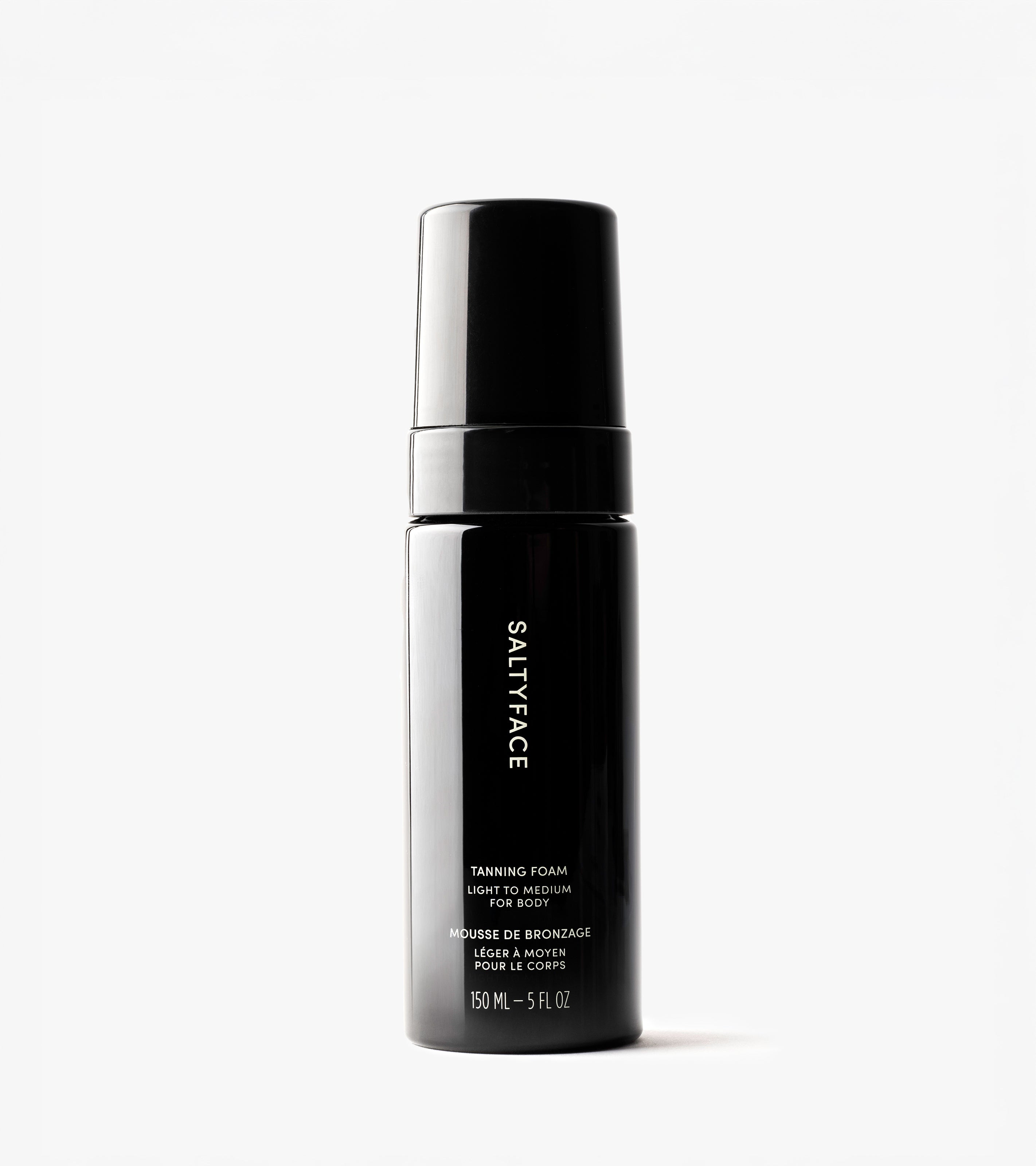 A black 150 mL bottle of Tanning Foam with a pump dispenser, labeled “Light to Medium” for face and body, features a water-to-foam formula for an easy, natural tan, and stands upright against a white background.