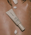 A tube of Tanning Moisturizer, celebrated for its gradual hydration, sits on sun-kissed skin amid sea shells and sprinkled sand.