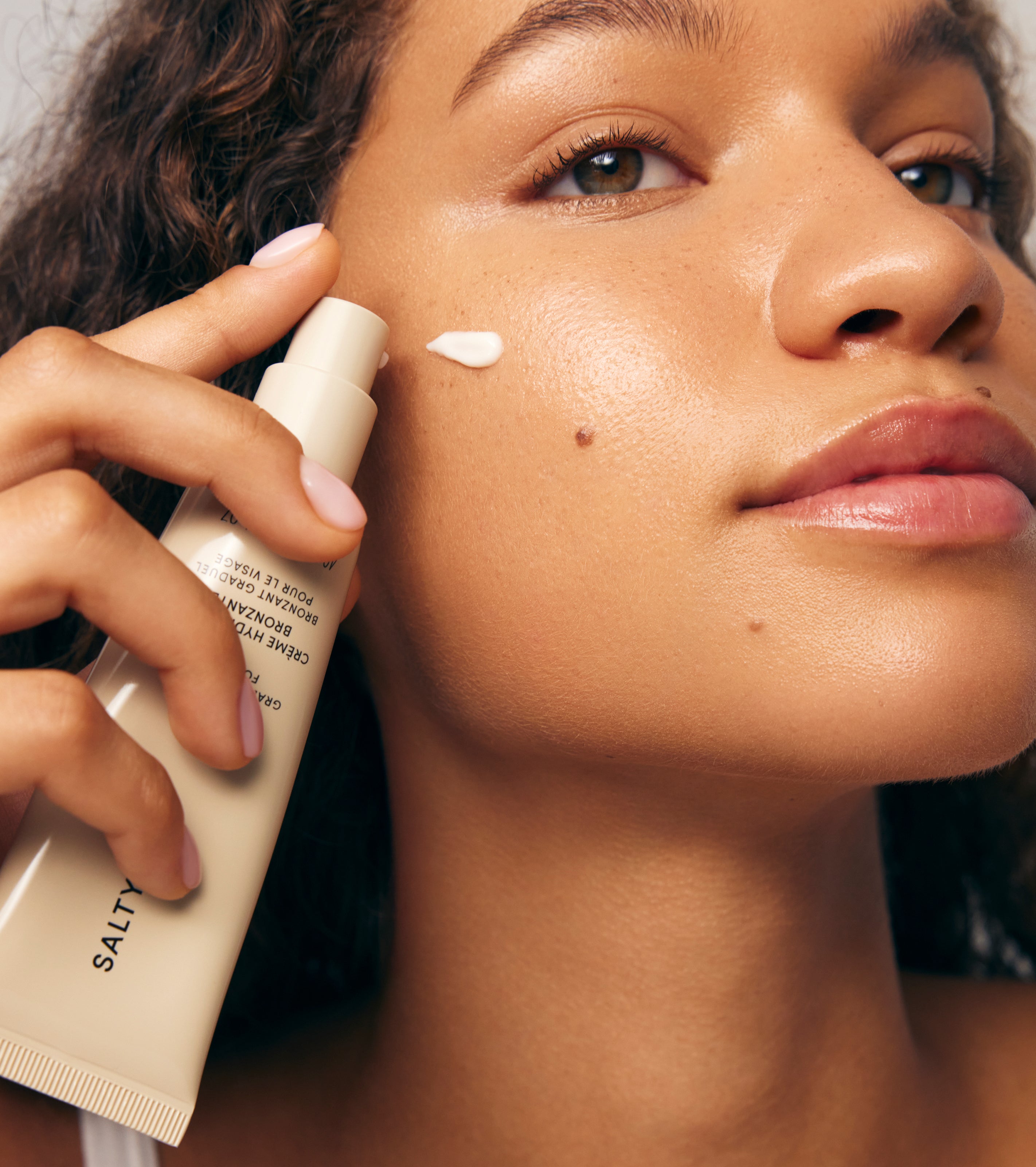 A woman applies Tanning Moisturizer to her cheek with her fingers, holding a beige tube labeled SALTY; her skin looks smooth, glowing, and subtly tanned.