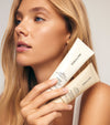 A woman with long blonde hair holds two beige The Moisturizer Duo tubes near her face, highlighting her radiant skin and natural makeup—ideal for achieving a sun-kissed look with The Moisturizer Duo.