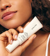 A person with curly hair holds a tube from The Moisturizer Duo near their face, highlighting smooth skin, natural makeup, and a touch of sun-kissed glow.