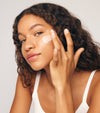 A woman with curly hair, in a white tank top, applies The Moisturizer Duo to her cheek with her fingers. She looks at the camera with a slight smile against a plain light background.