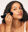 A woman with curly hair applies Liquid Bronzer to her cheek using Brush No. 3, a black double-ended brush. She wears a white tank top and has clear, glowing skin with a calm expression, demonstrating seamless application.