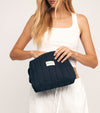A woman in a white crop top and shorts holds The Everywhere Bag, a black quilted zip pouch with a SALTYFACE label, opening it with one hand against a plain light background—ideal as an essentials organizer for daily use.