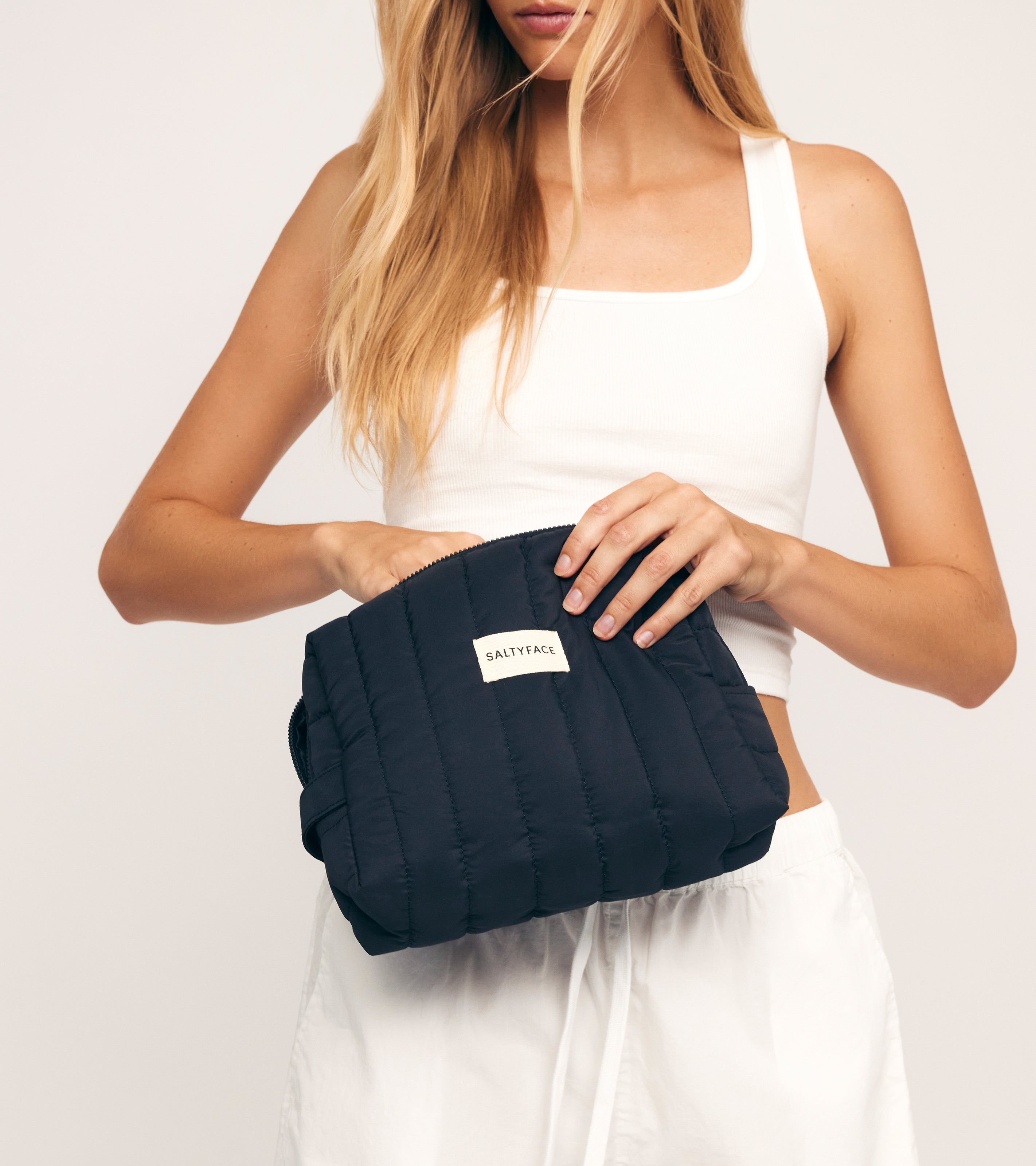 A woman in a white crop top and shorts holds The Everywhere Bag, a black quilted zip pouch with a SALTYFACE label, opening it with one hand against a plain light background—ideal as an essentials organizer for daily use.