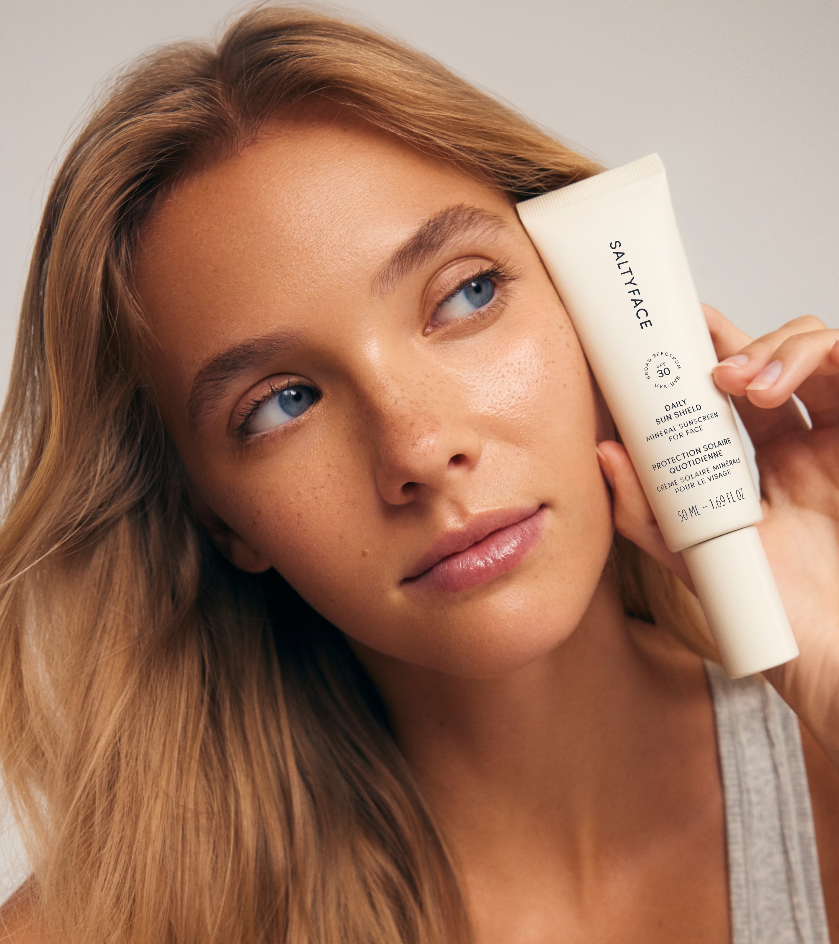 A woman with long blonde hair holds a tube of Daily Sun Shield close to her face, looking slightly to the side with a calm expression against a softly lit, neutral background.