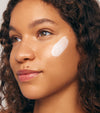 A woman with curly hair and glowing skin wears a swipe of Daily Sun Shield SPF 30 cream on her cheek, looking to the side with a calm expression to highlight the weightless feel of the sunscreen.