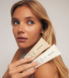 A woman with long blonde hair holds two beige Sunscreen & Tan tubes near her face, looking to the side against a plain background, emphasizing the importance of hydrated skin and sun protection in her beauty routine.