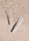 Two beige Sunscreen & Tan tubes rest on sand beside clear crystals. Sunlight highlights their clean design, hinting these Salt & Stone essentials deliver mineral sun protection and a radiant glow for sun-kissed skin.