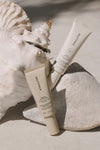 Two Sunscreen & Tan tubes rest on pale rocks beside a large seashell, with natural sunlight highlighting their minimalist packaging against a neutral background.
