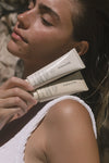 A woman with sun-kissed skin in a white textured top holds two Sunscreen & Tan tubes near her face, eyes closed, enjoying the sunlight. The softly blurred background enhances the serene moment.