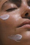 A close-up of a person’s face with eyes closed, revealing dewy skin and three patches of Sunscreen & Tan SPF 30 mineral cream applied on the cheek and chin.