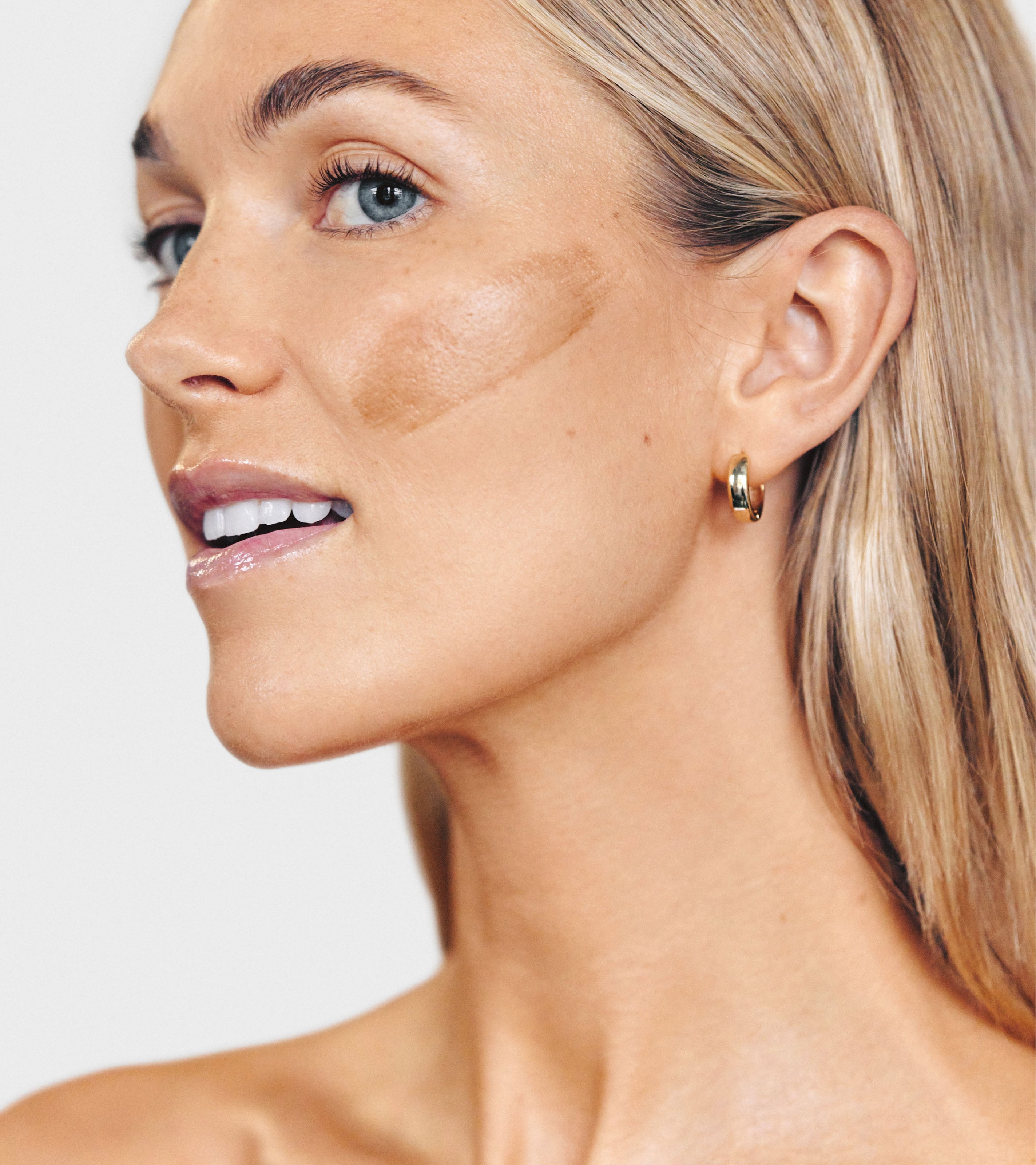 A woman with light skin and blonde hair wears small hoop earrings and applies a swipe of brown foundation on her cheek using Brush No. 3, highlighting her flawless complexion as she smiles softly in profile.