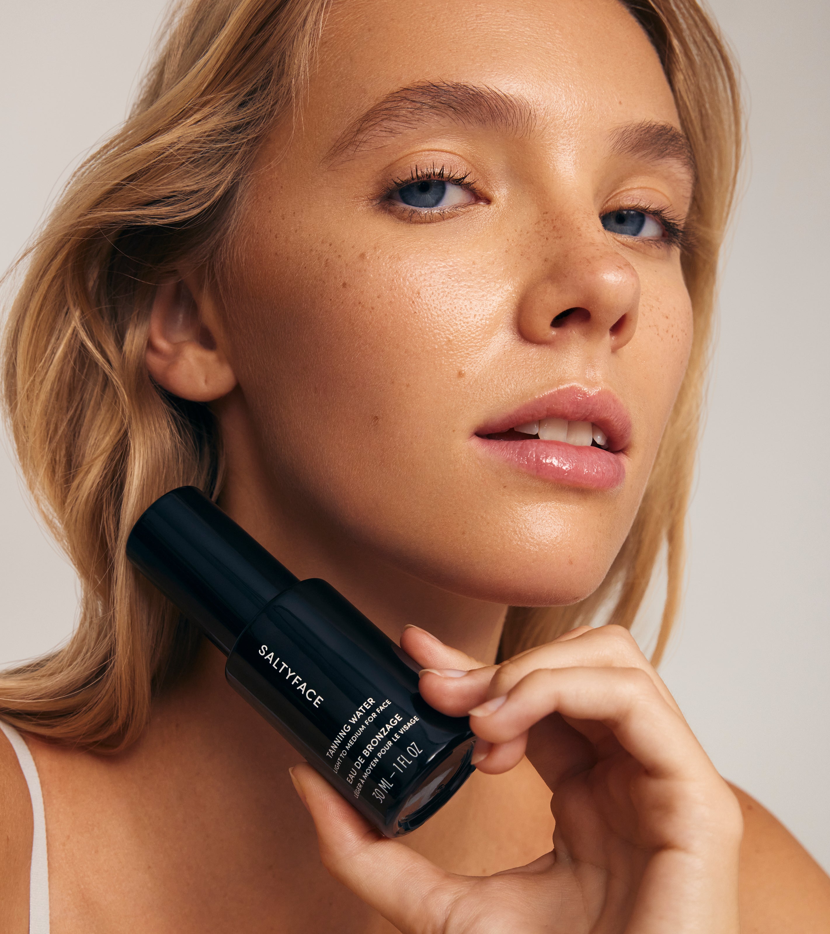 A woman with blonde hair and clear skin confidently holds a black bottle of Tanning Water near her chin, her sun-kissed tan and radiant glow enhanced by the softly lit, neutral background.