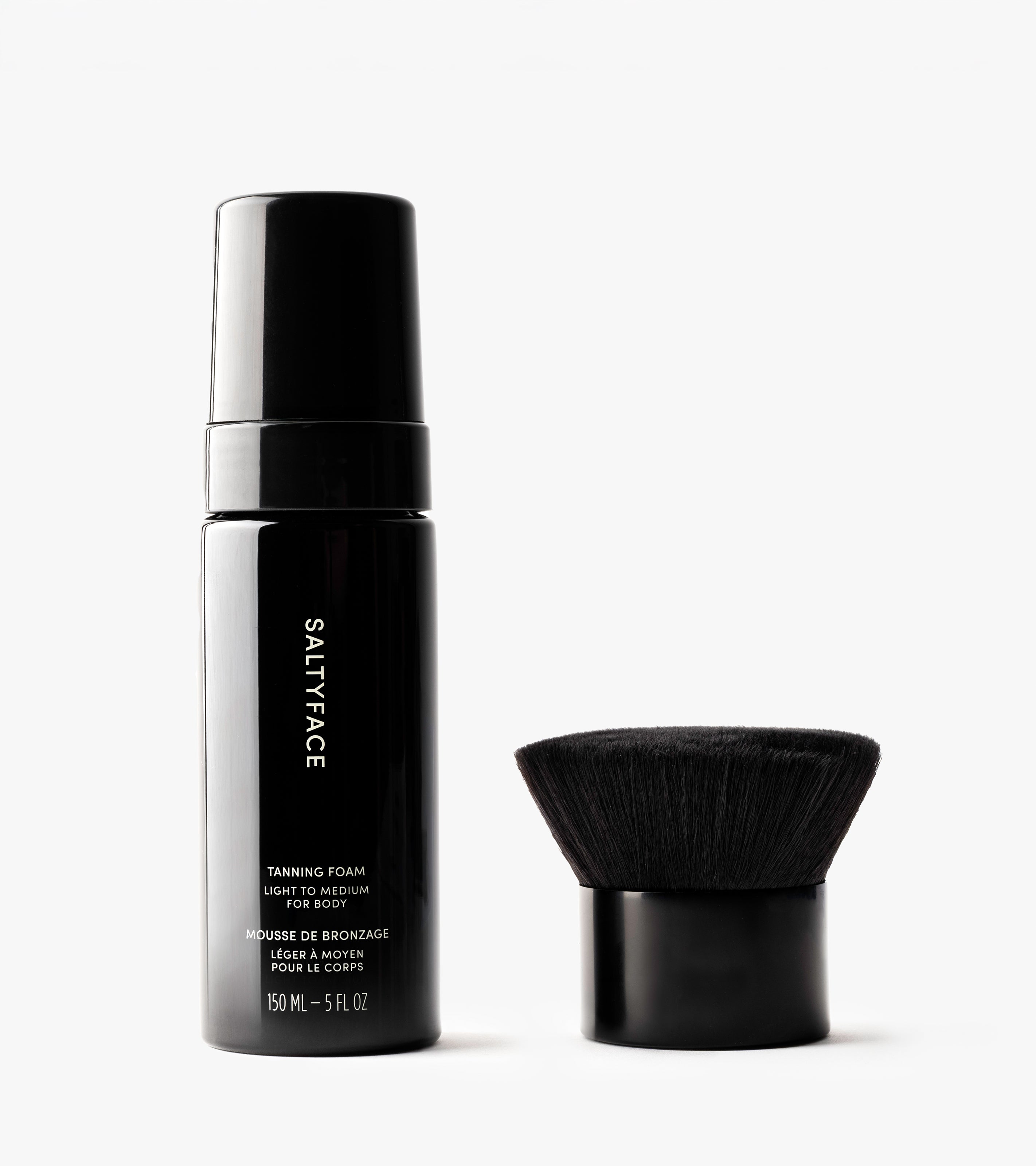 A black bottle from The Body Set stands beside a short, dense body brush on a white background. The bottle features white text and a pump top, ideal for achieving a flawless, sun-kissed tan.