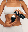 A woman in a light grey sports bra and shorts dispenses The Body Set Tanning Foam from a black bottle onto a black body brush, holding both before her bare midriff for a flawless, sun-kissed tan.