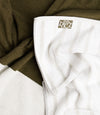 The Summer Towel, white with light gray stripes and a PLAYA label, is slightly crumpled and partially overlaps atop textured olive-green fabric.