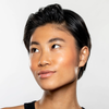 A person with short, dark hair and a gold septum ring looks upward with a slight smile, wearing a black top. Their smooth, glowing skin showcases the radiance of Gen See – Clean Sheen against a plain, light-colored background.