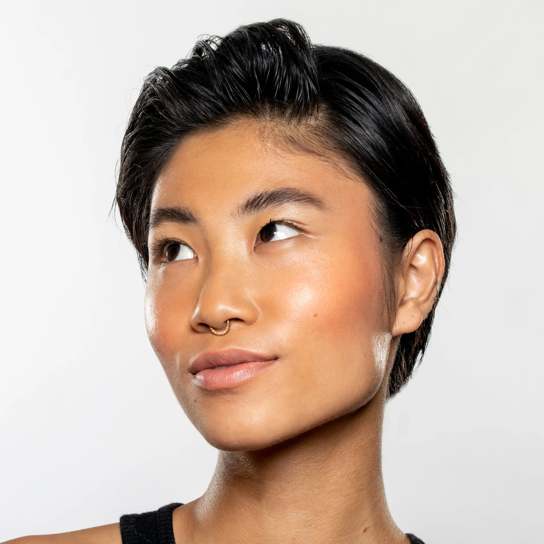 A person with short, dark hair and a gold septum ring looks upward with a slight smile, wearing a black top. Their smooth, glowing skin showcases the radiance of Gen See – Clean Sheen against a plain, light-colored background.