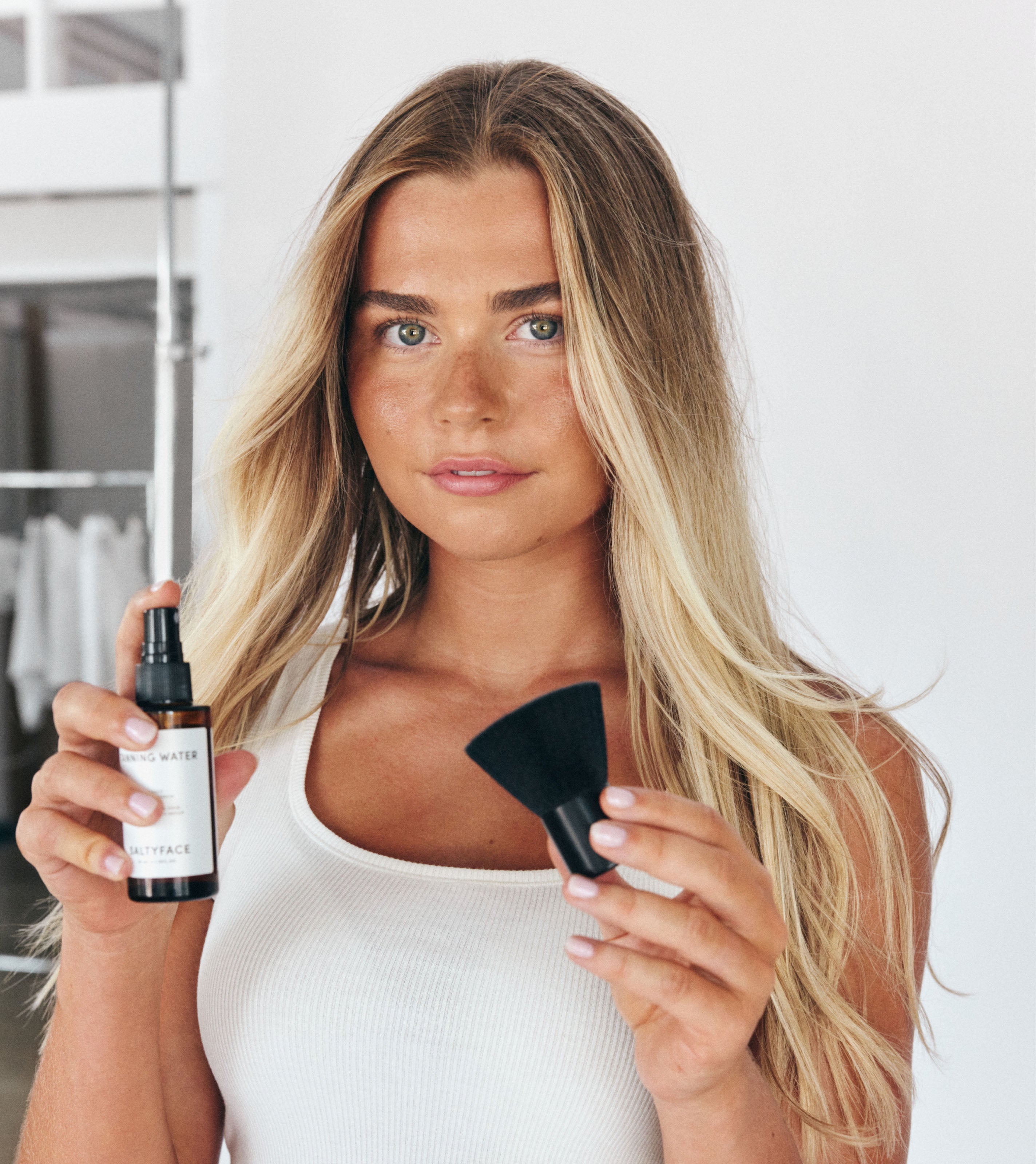 Model holding Saltyface Tanning Water with a Mist Top in one hand, and Brush No. 2 in the other.