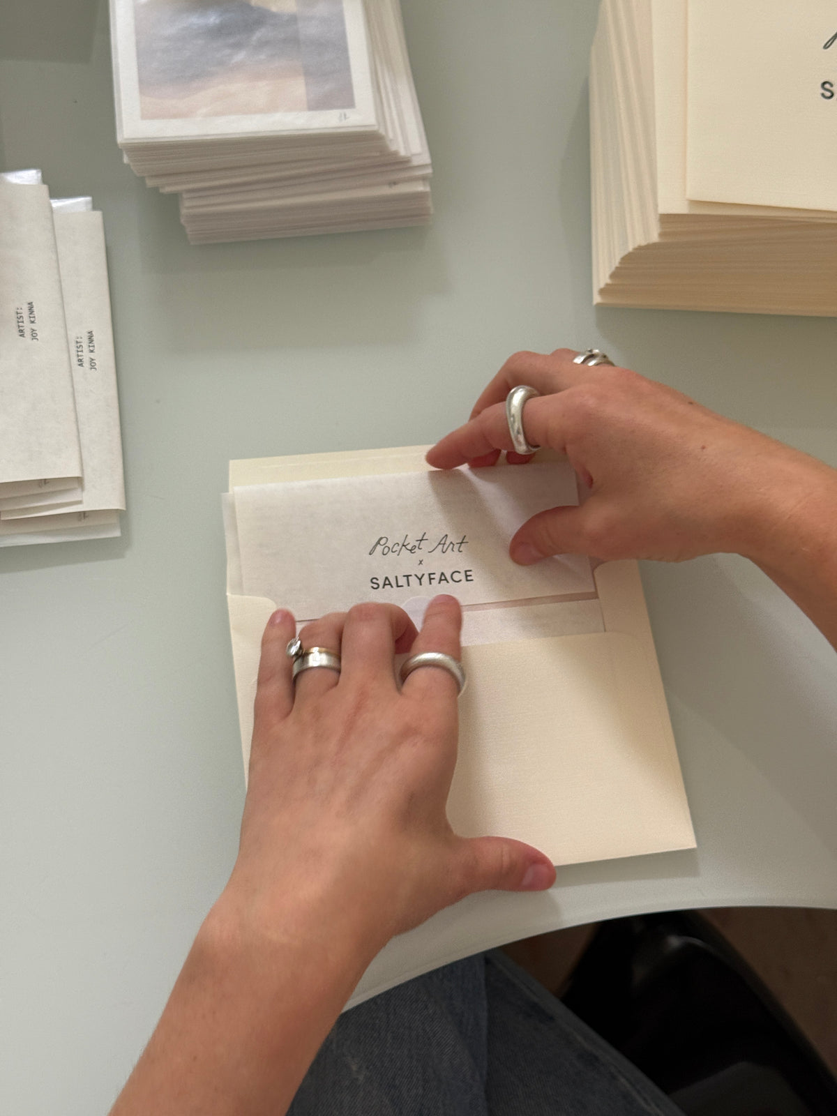 Hands with silver rings insert a Pocket Art card into an envelope on a white table, surrounded by stacks of paper and envelopes.