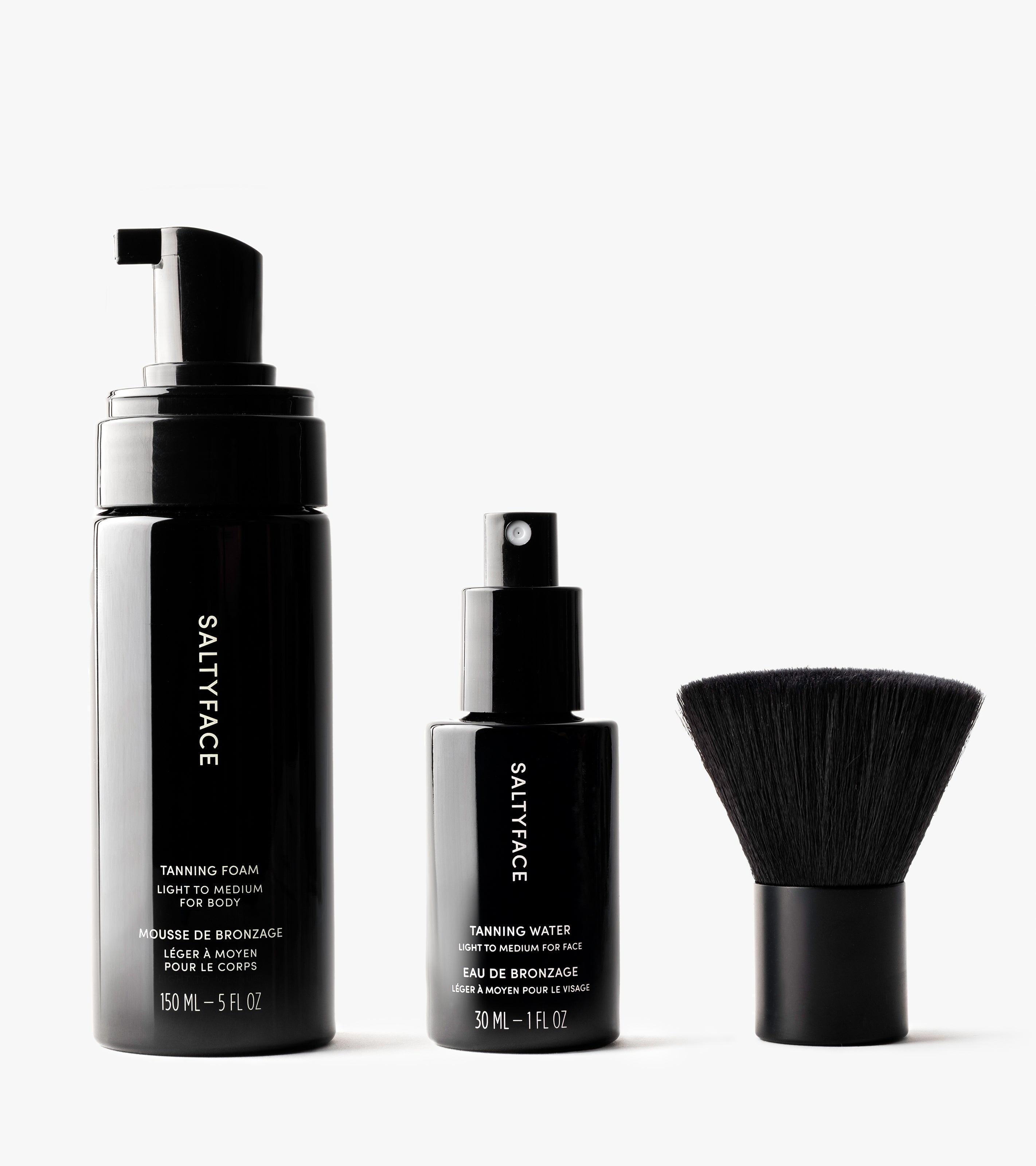 The Essential Face & Body Set is displayed upright on a white background, featuring three black SALTYFACE products: a Tanning Foam bottle with pump, a smaller Tanning Water spray bottle, and a short, wide face brush.