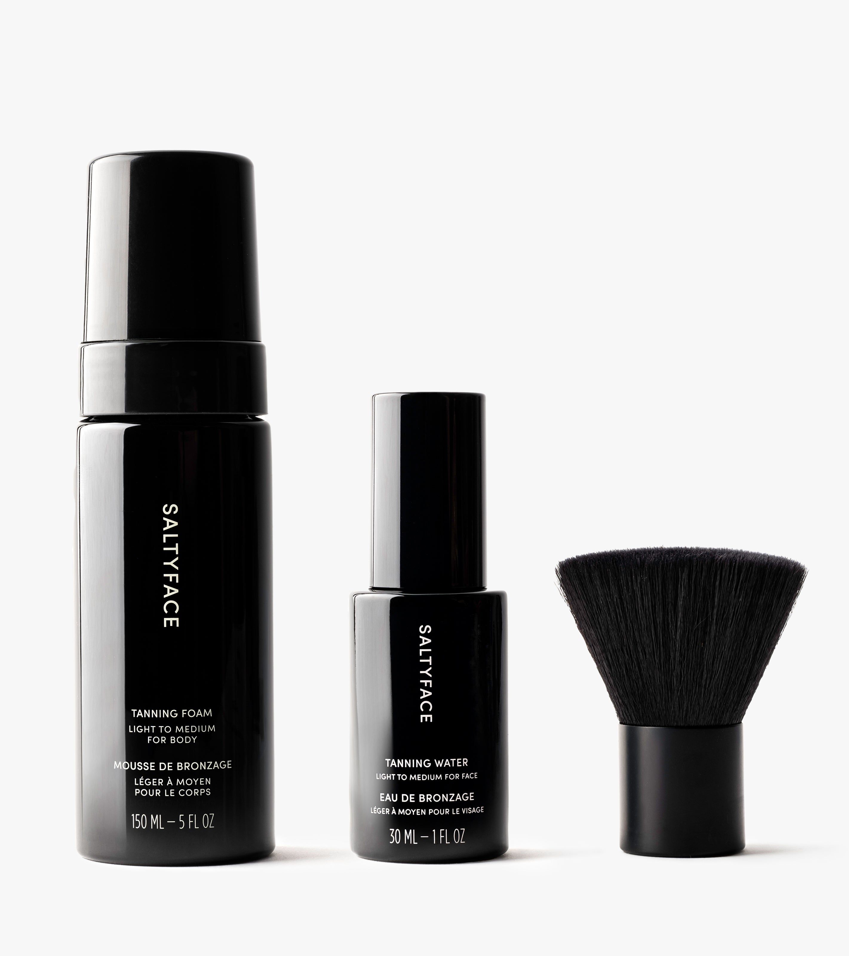 The Essential Face & Body Set is shown with three black products—a 150 mL Tanning Foam, a 30 mL Tanning Water, and a short round face brush—standing upright on a white background.