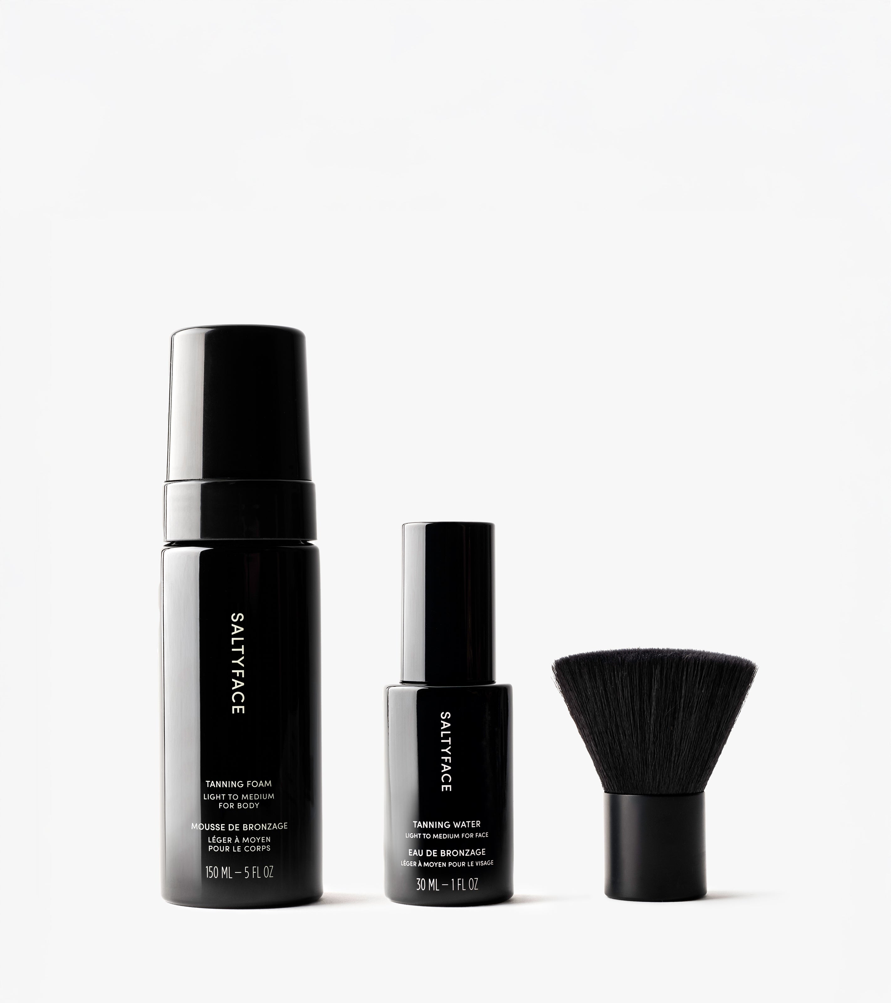 The Essential Face & Body Set features three black cosmetic products—a tanning foam, tanning water, and a short, dense-bristled face brush—displayed on a white background.