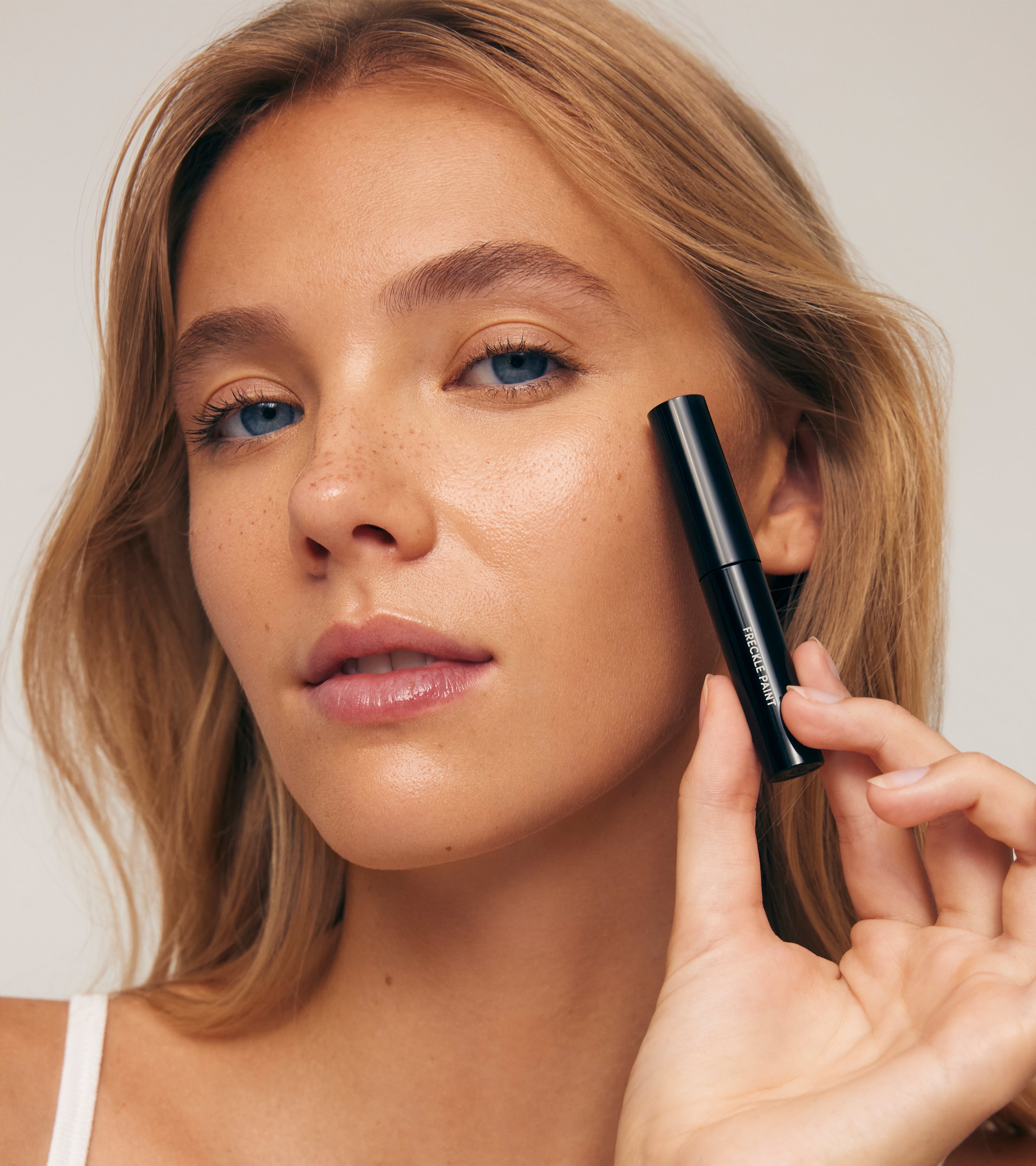 A woman with light skin and natural freckles confidently holds a tube of Freckle Paint near her cheek. She wears natural makeup, a white top, and looks directly at the camera.