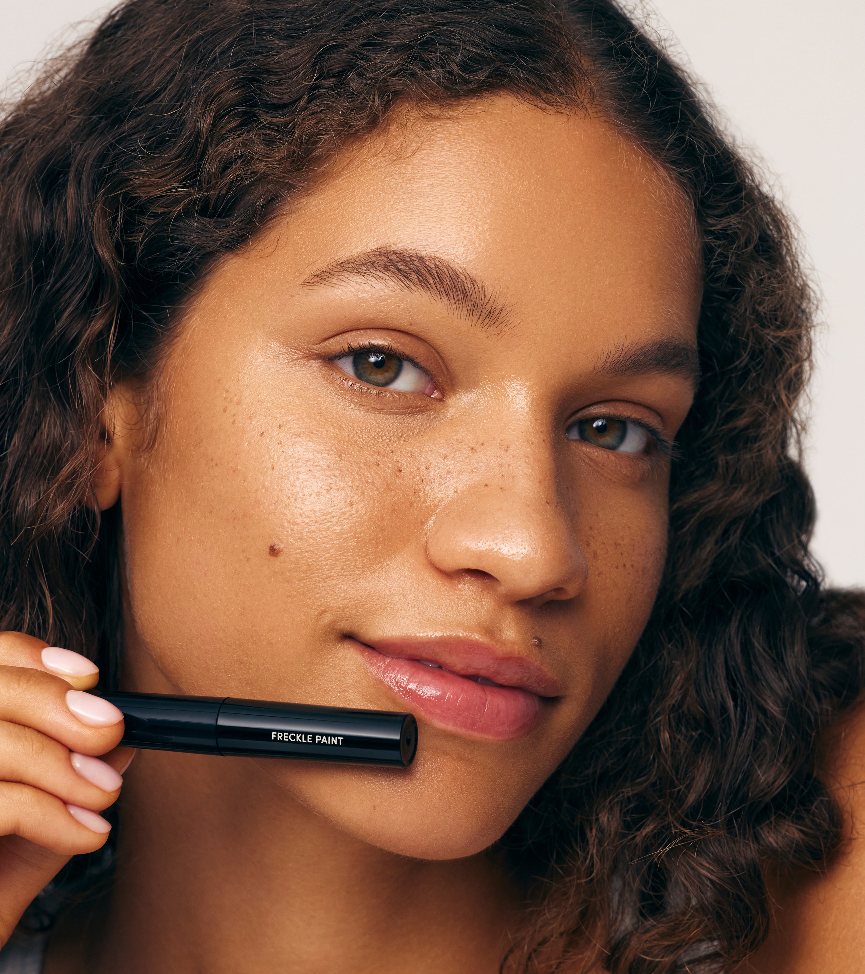 A woman with curly hair and natural makeup holds a tube of Freckle Paint - REBRAND near her face, showing off subtle, long-lasting freckles and smooth, glowing, sun-kissed skin.