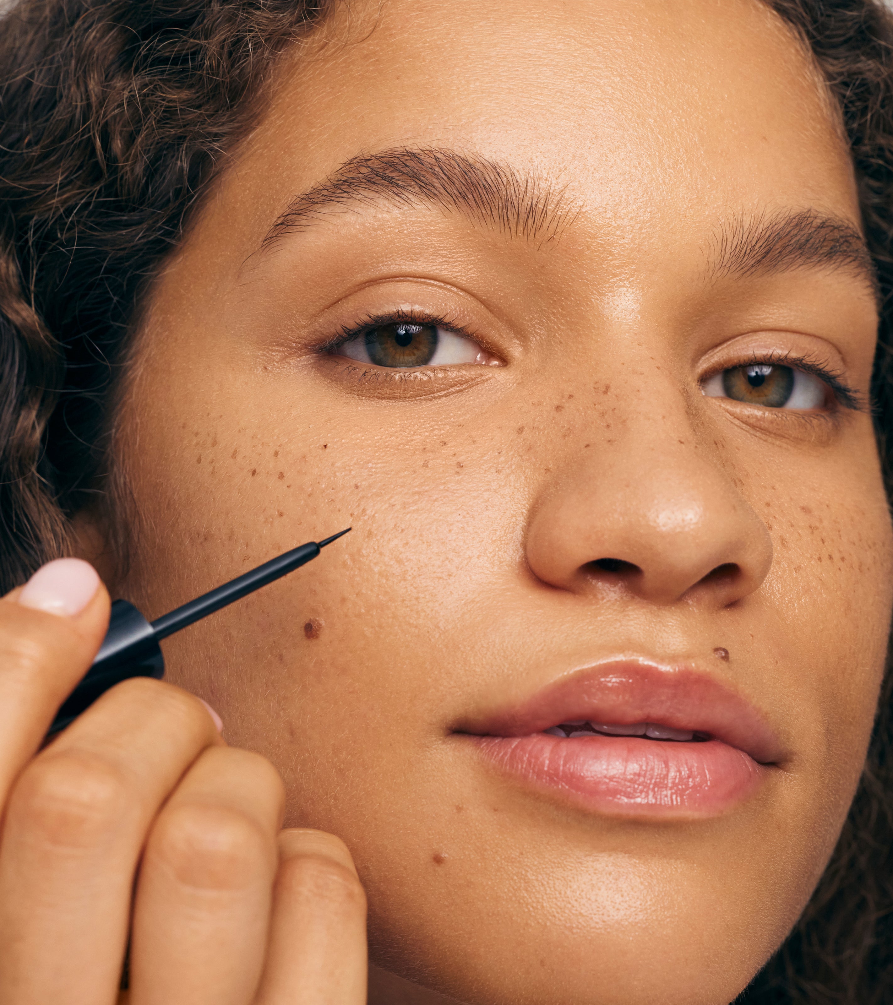 A person with curly hair and natural freckles holds Freckle Paint near their cheek. Their glowing skin, glossy lips, and radiant, sun-kissed makeup highlight a fresh, natural look.