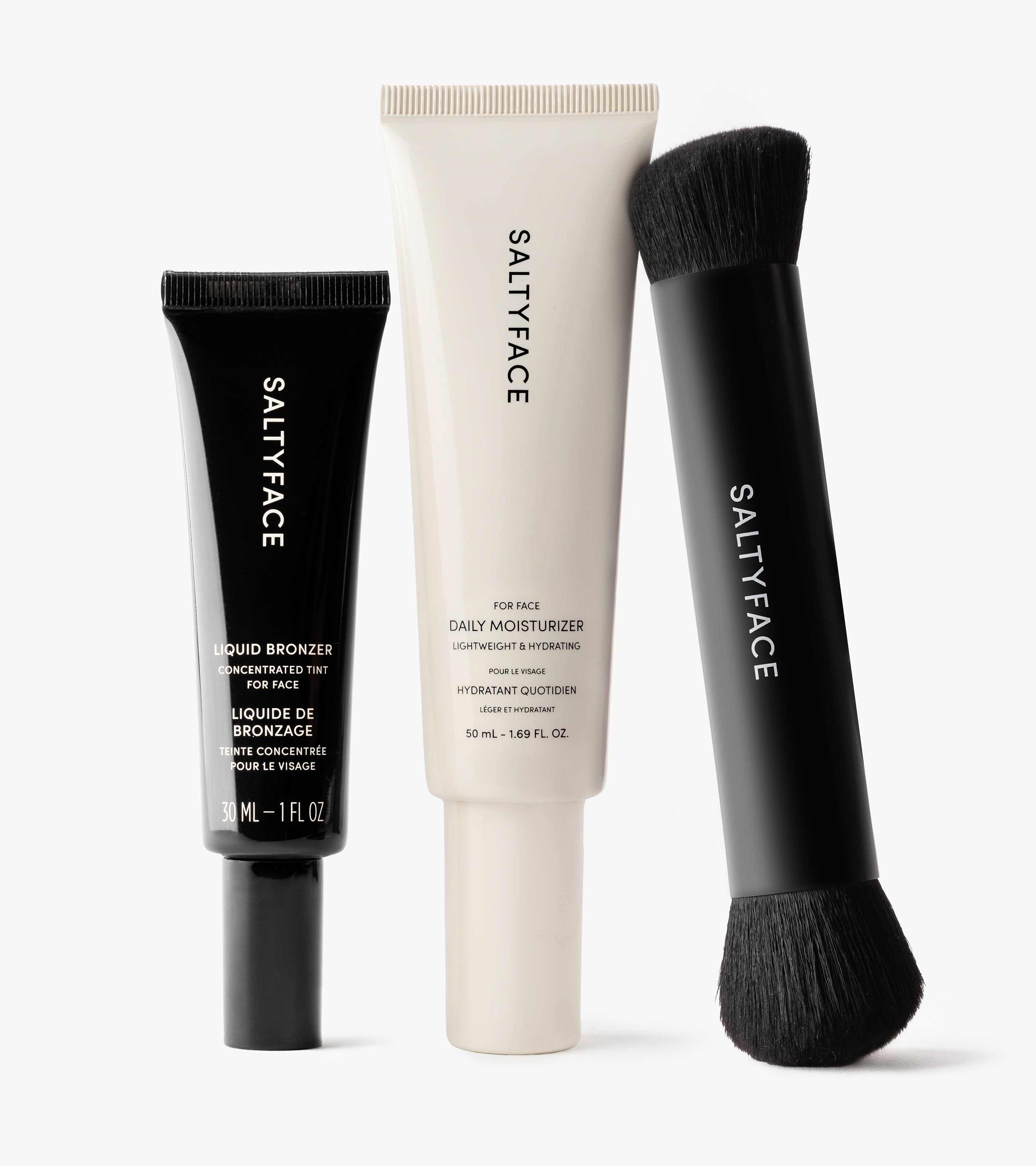 The Hydrate & Bronze Set features a black tube of Liquid Bronzer for a natural contour and instant glow, a white tube of daily moisturizer, and a dual-ended black makeup brush, all standing upright against a white background.
