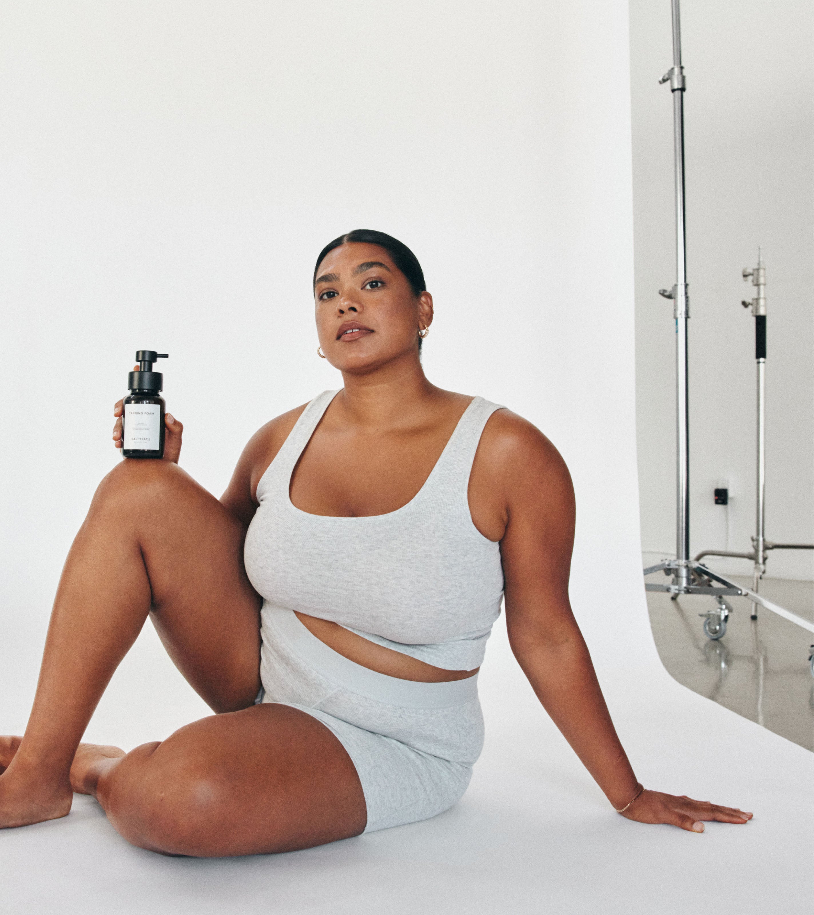 Model in a grey lounge set holding Saltyface Tanning Foam with a tanned complexion. 