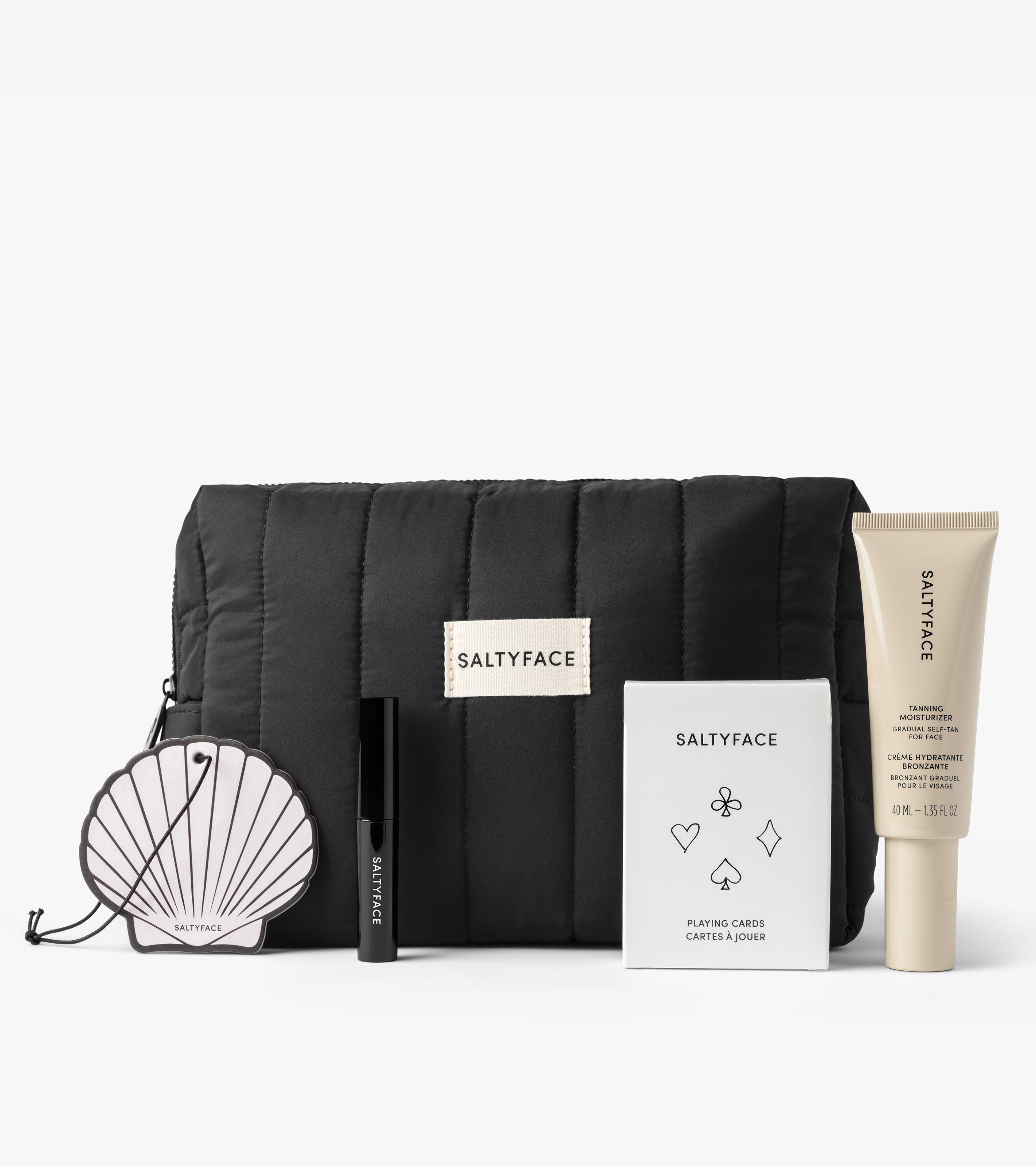 The Road Trip Set features a black quilted SALTYFACE pouch, a shell-shaped mirror, serum tube, mascara, and a deck of playing cards, all neatly arranged against a white background.