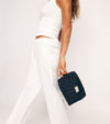 A person in a white sleeveless top and pants holds a small black quilted bag, perfect for carrying essentials like The Holiday Face Set, against a plain background.
