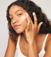 A woman with curly hair applies The Holiday Duo to her cheek with her fingers. She is wearing a white tank top and looking at the camera against a neutral background—perfect for glowing skin before holiday travel.