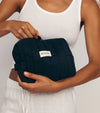 A person in a white tank top and pants holds The Holiday Duo—a black quilted pouch with a zipper and SALTYFACE label—ideal for holiday travel or storing your gradual tanning moisturizer.
