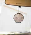 A “The Sun Set” air freshener in the shape of a shell hangs from a car’s rearview mirror. The softly blurred background reveals a light-colored building outside the vehicle.
