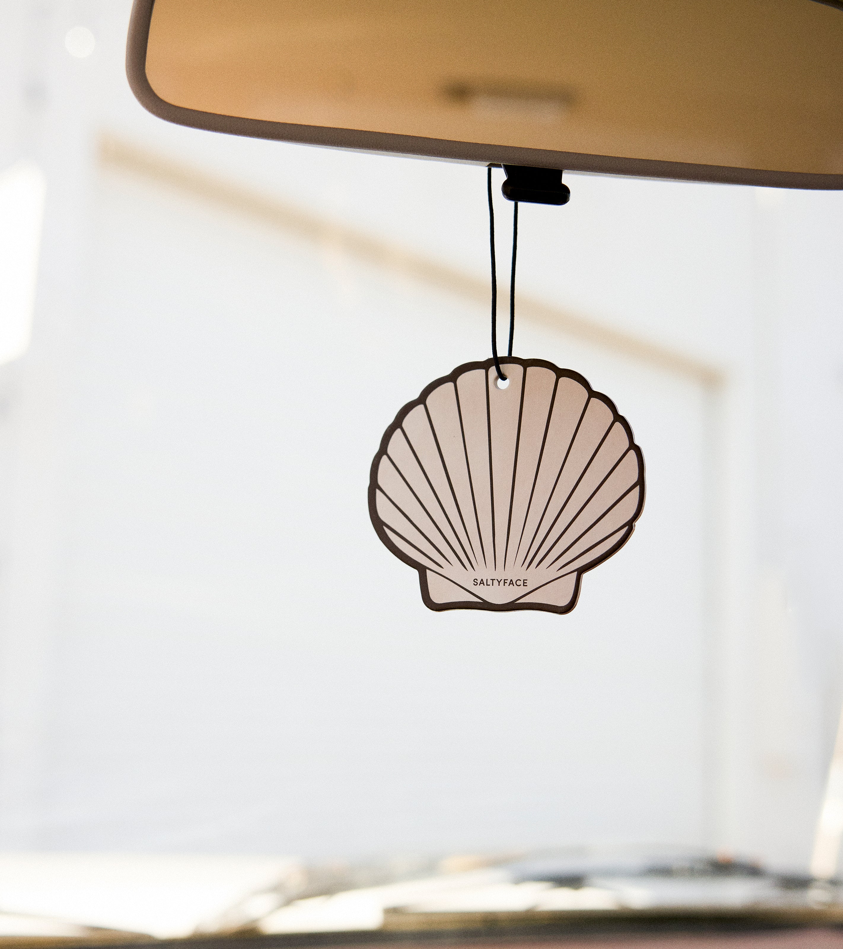 A “The Sun Set” air freshener in the shape of a shell hangs from a car’s rearview mirror. The softly blurred background reveals a light-colored building outside the vehicle.