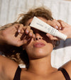 A woman with wavy hair lies on a towel in the sunlight, covering one eye with her hand and holding up The Sun Set skincare tube, looking relaxed.