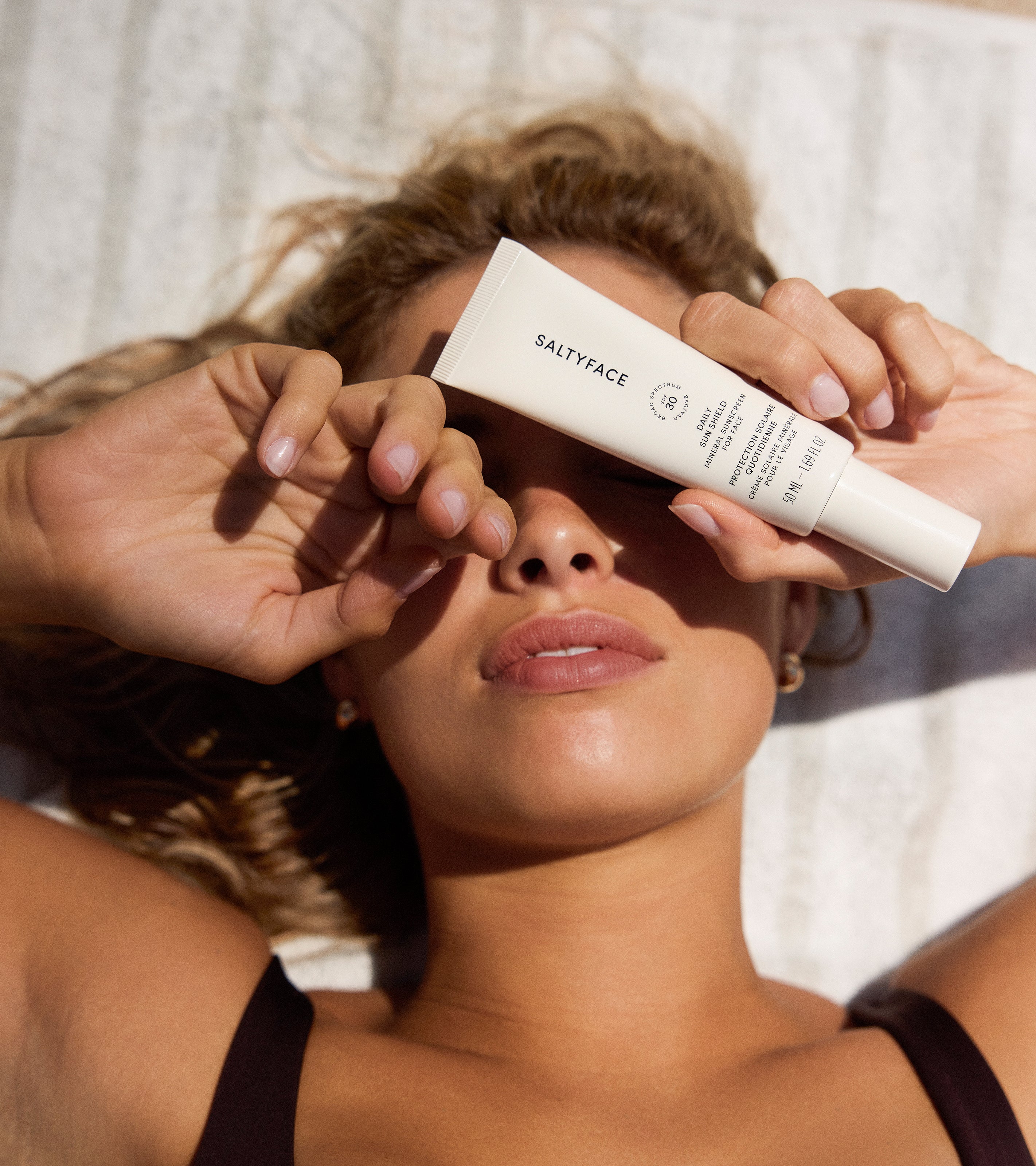 A woman with wavy hair lies on a towel in the sunlight, covering one eye with her hand and holding up The Sun Set skincare tube, looking relaxed.
