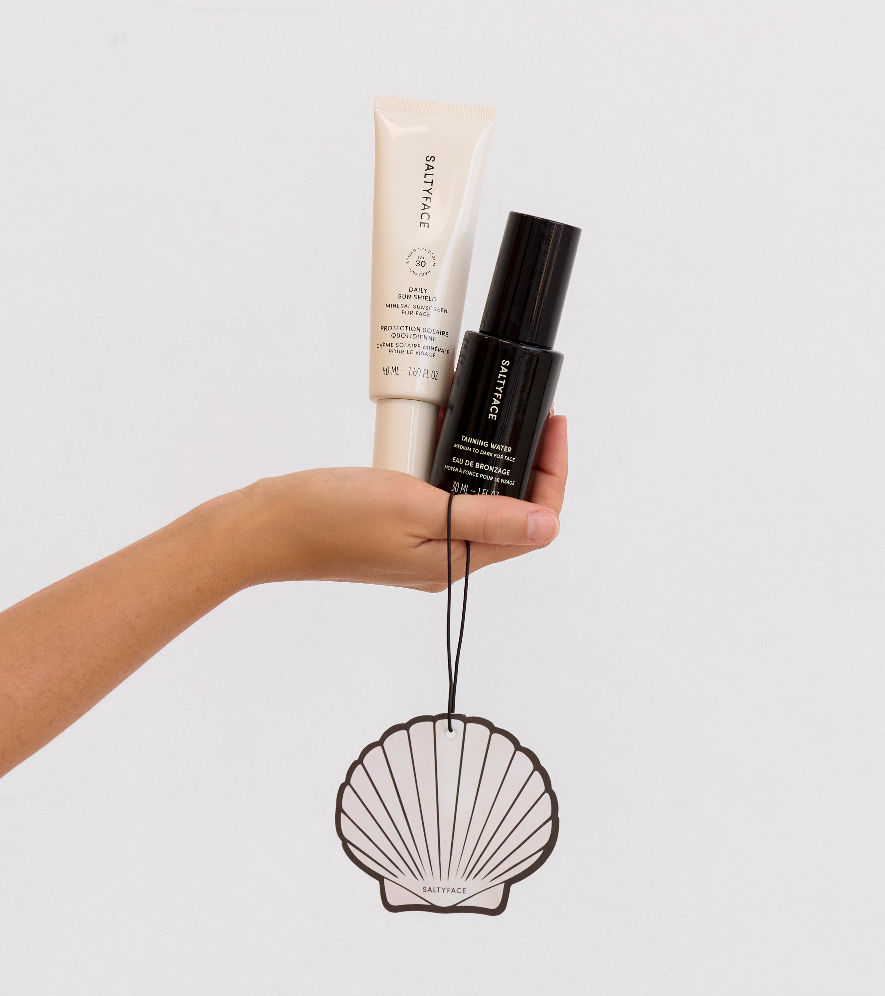 A hand holds The Sun Set products—a white tube, a black spray bottle, and a shell-shaped hanging decoration—against a plain white background.