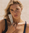 A woman with long blonde hair and freckles holds The Road Trip Set skincare tube near her face. She wears a dark tank top and stands outside in natural light.