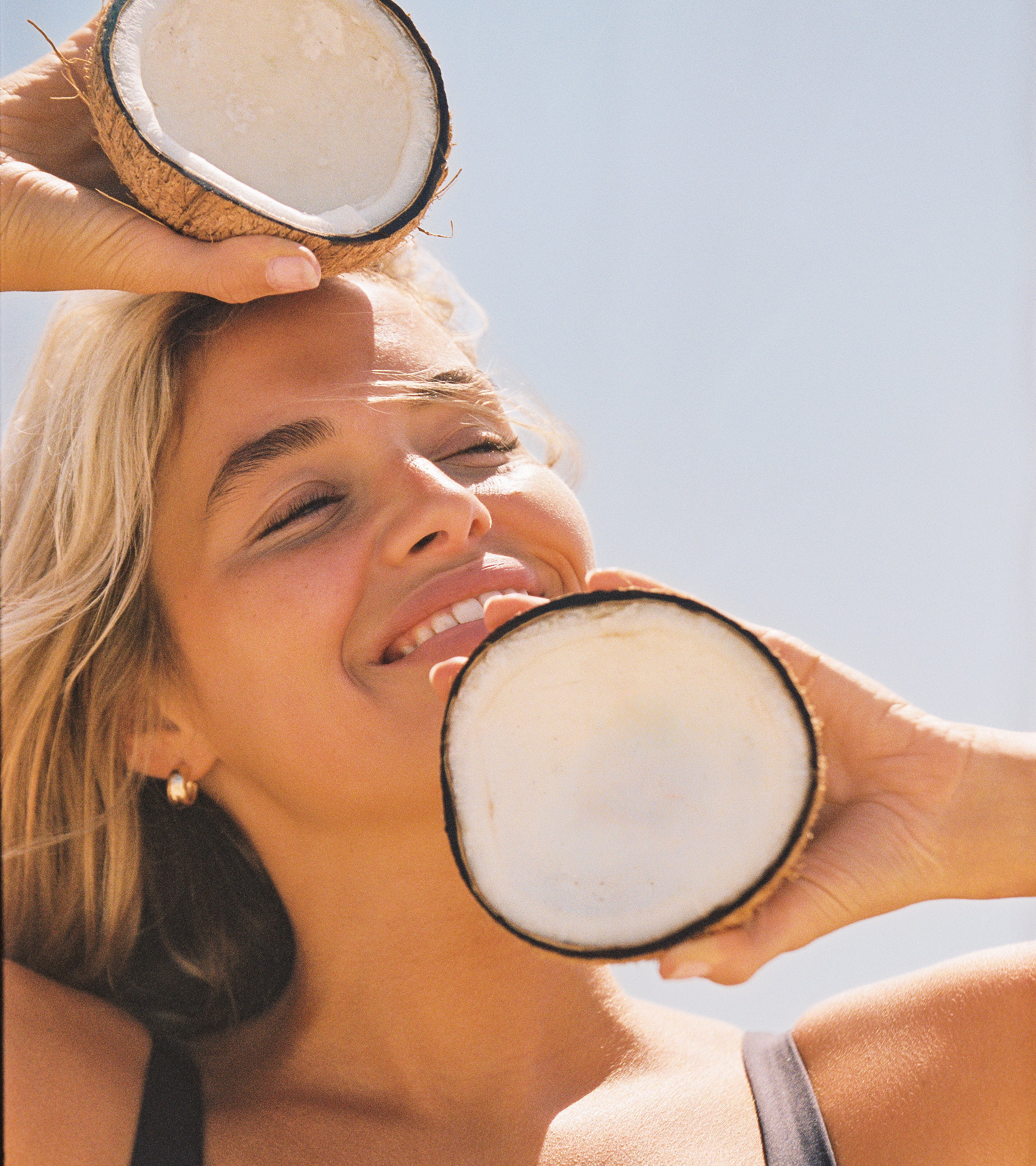 A smiling person holds two coconut halves—one above their head, one near their chin—beneath a clear blue sky, evoking the fresh, tropical scent of Air Freshener. Sunlight highlights their face and hair for a vibrant scene.