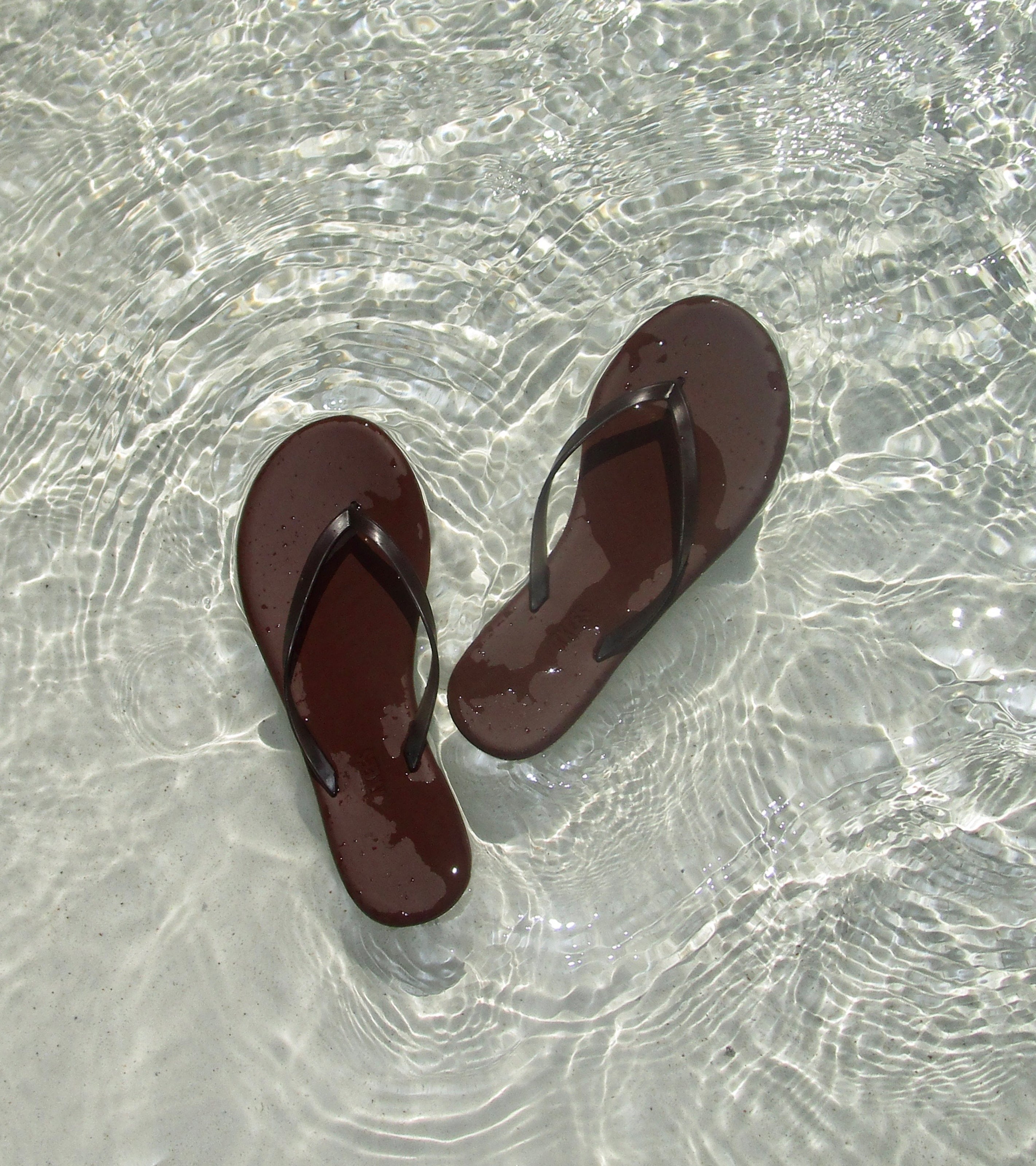 Aqua Flip Flops in dark brown float in clear, shallow water as sunlight creates gentle ripples and shimmering reflections on the sandy bottom.