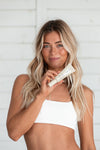 A smiling woman with long wavy blonde hair in a white bandeau top holds up a beige tube of Tanning Moisturizer, showcasing her glowing gradual tan against a white wooden wall.