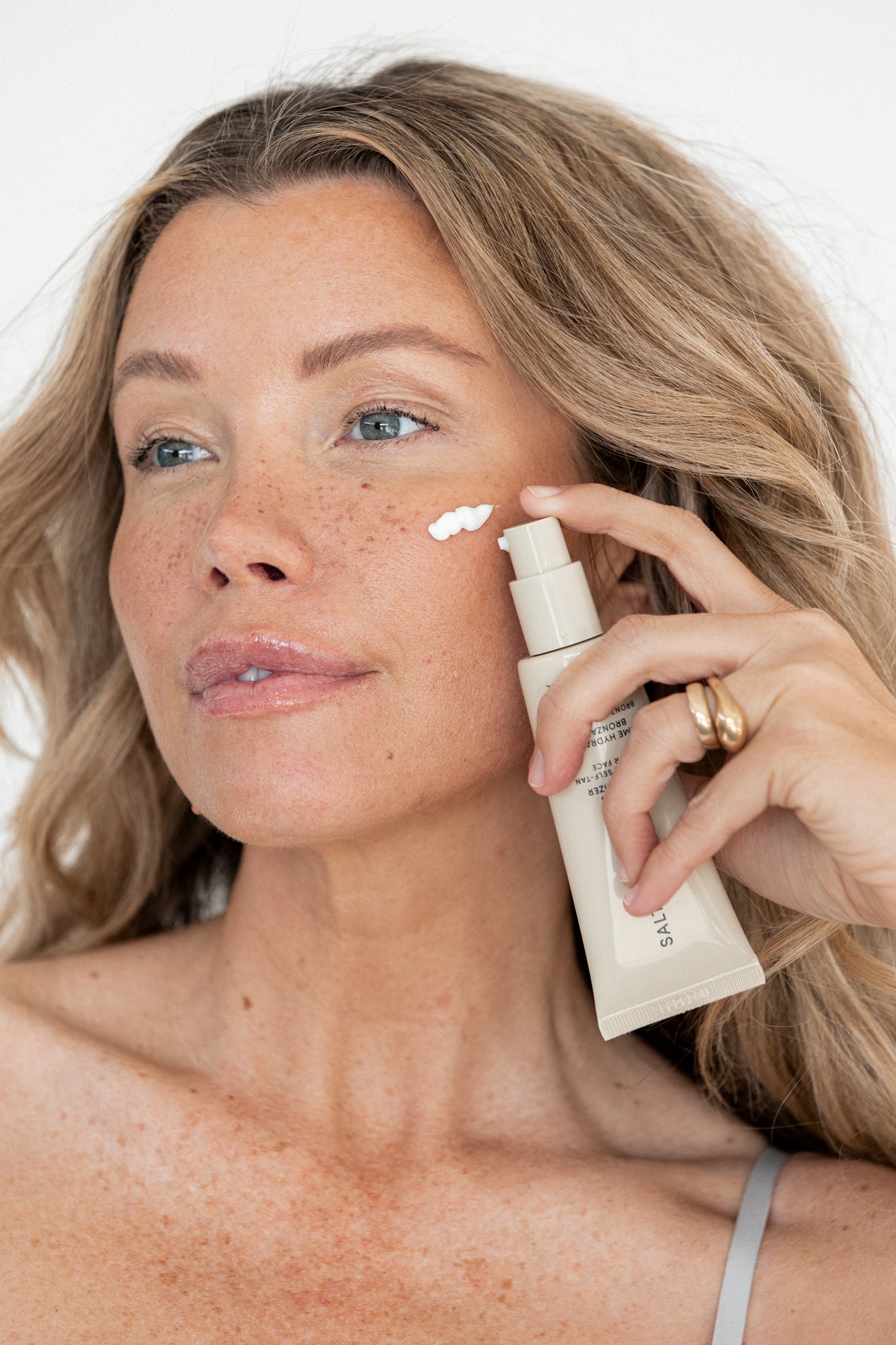 A woman with wavy light brown hair applies The Moisturizer Duo to her cheek, holding its beige tube. She wears a gold ring and thin strap top, smiling softly against a plain background.