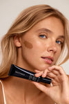 A light-skinned, blonde woman holds a tube of Liquid Bronzer v2 labeled Saltyface to her face, displaying a streak of the bronzer on her cheek to demonstrate its makeup application.